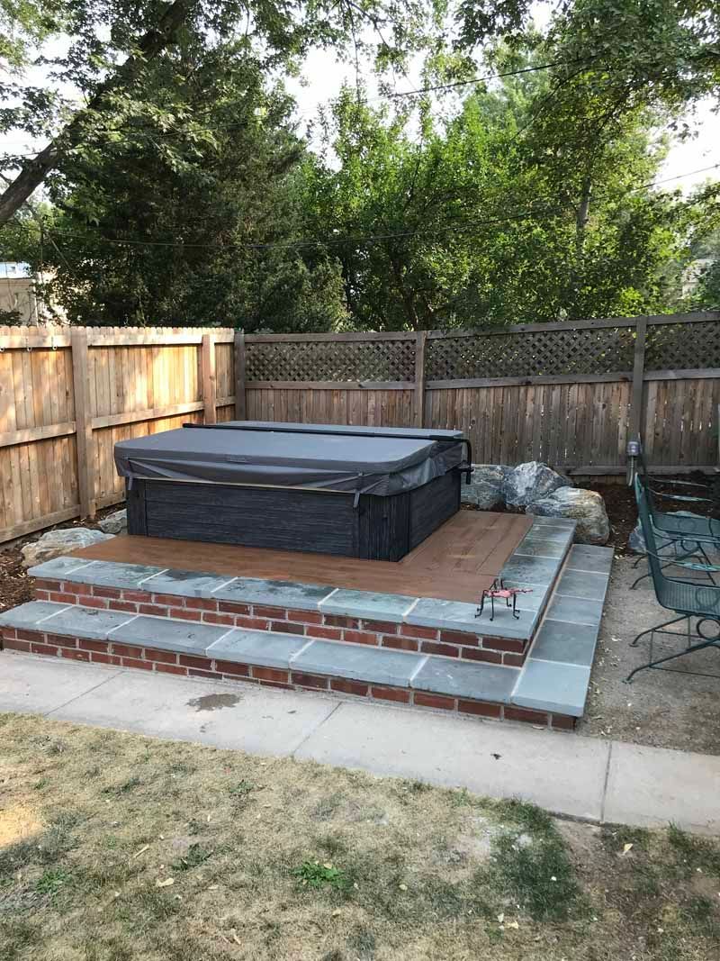 A dark gray hot tub with a cover sits on a tiered brick and stone patio in a backyard with a wooden fence.