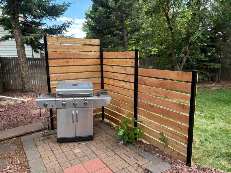 A modern wooden slat privacy screen with a stainless steel gas grill on a brick patio in a backyard setting.