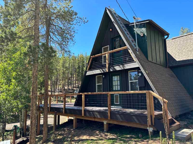 A dark-paneled, A-frame mountain cabin with a large wooden deck and balcony, nestled among evergreen trees.