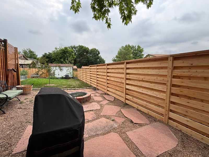 A backyard scene with a modern horizontal wood fence, a stone patio with a fire pit, and a covered grill on gravel.