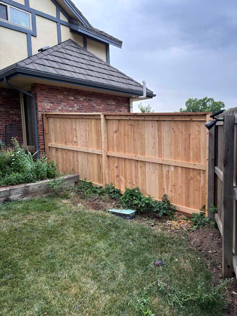 A new cedar wood fence stands in a grassy backyard next to the brick wall of a house under a cloudy sky.