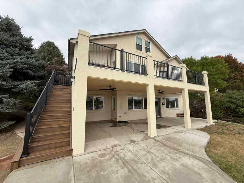 A beige, two-story house with a wooden staircase leading to a large upper deck and a concrete patio on the ground level.