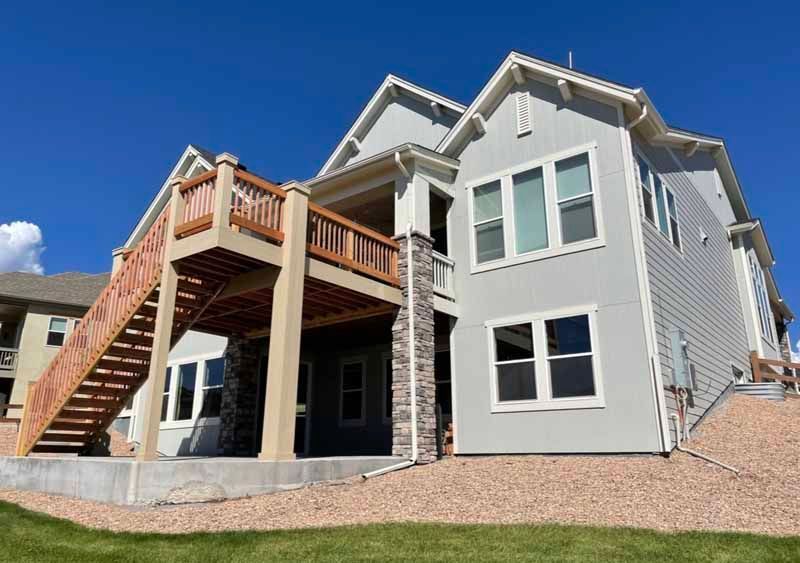 A light gray two-story house with stone accents, featuring a large wooden deck with stairs leading to a concrete patio.