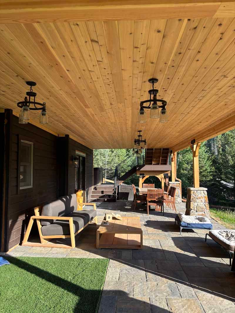 Covered wooden patio with comfortable outdoor seating, a stone floor, and pendant lights overlooking a wooded area.