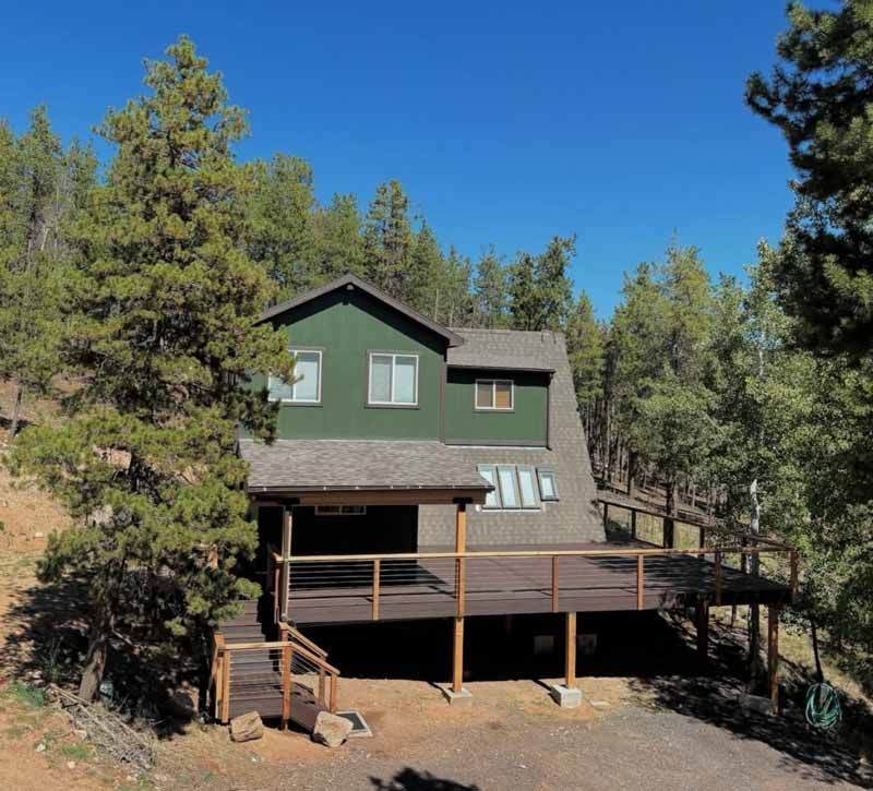 A green two-story mountain home with a large wooden deck, nestled among evergreen trees under a clear blue sky.