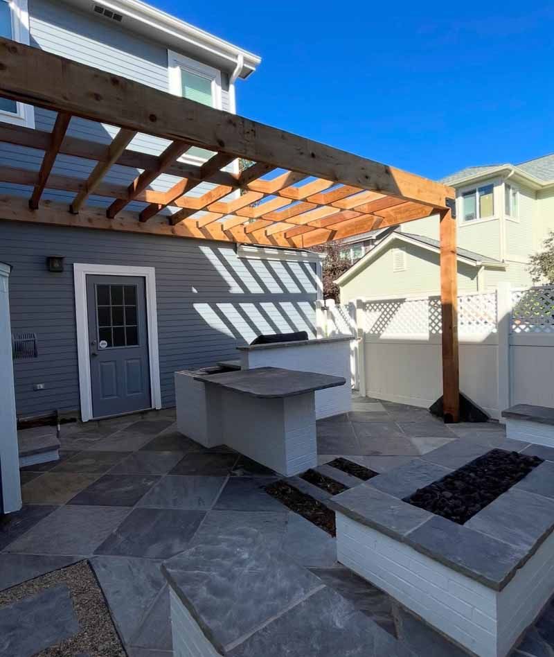 A gray house patio with a wooden pergola, a built-in outdoor kitchen island, and a stone fire pit under a clear blue sky.