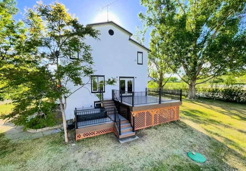 A two-story white house with a multi-level wooden deck featuring black railings and lattice skirting, set in a yard.
