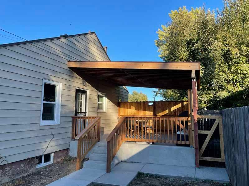 A light-colored house exterior featuring a wooden deck with a covered patio, stairs, and a concrete walkway.