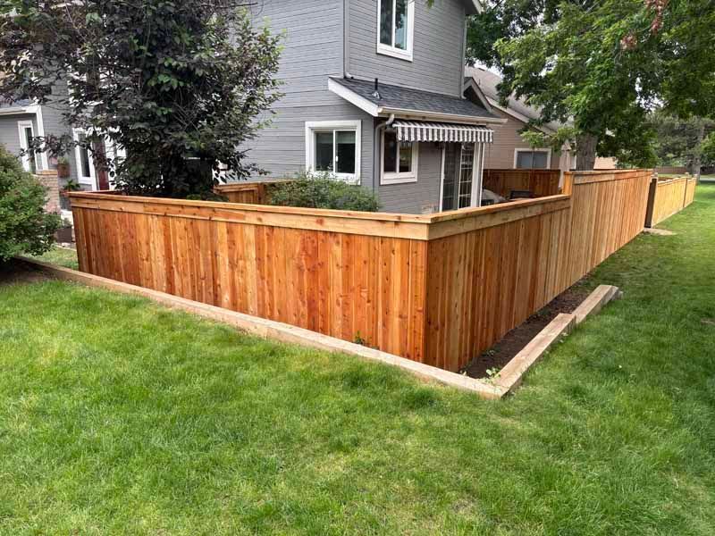 A new, stained cedar wood privacy fence borders a grassy yard in front of a gray two-story house.