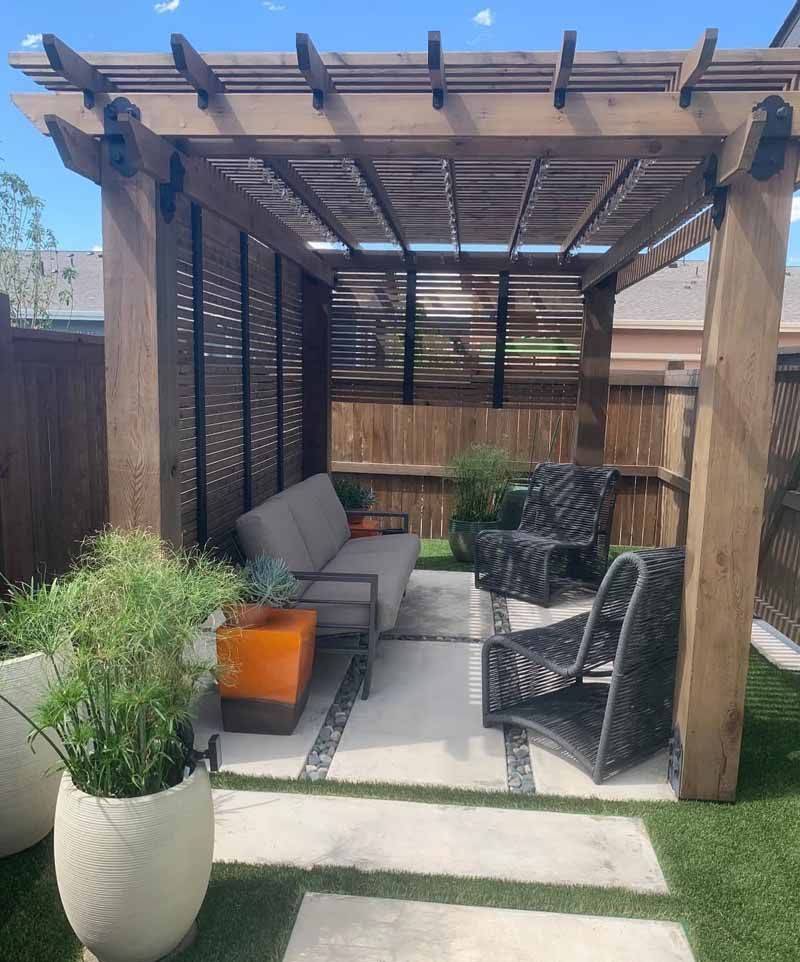 A wooden pergola patio with an outdoor sofa, two woven chairs, an orange side table, and potted plants on stone pavers.