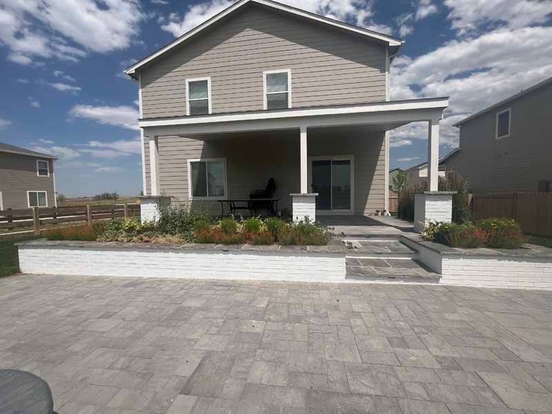 A tan two-story house with a white-walled patio area, stone pavers, and landscaping under a blue sky with light clouds.