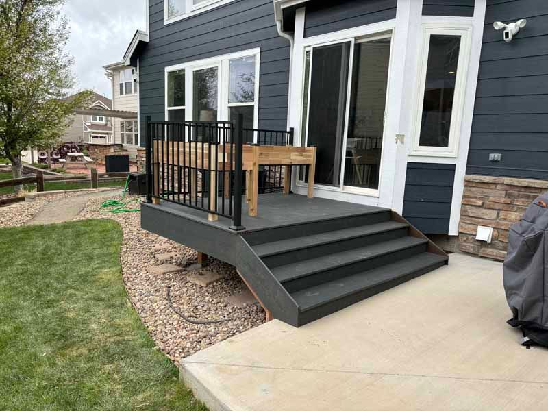 A dark gray deck with black metal railings and stairs leading down to a concrete patio outside a dark-sided house.