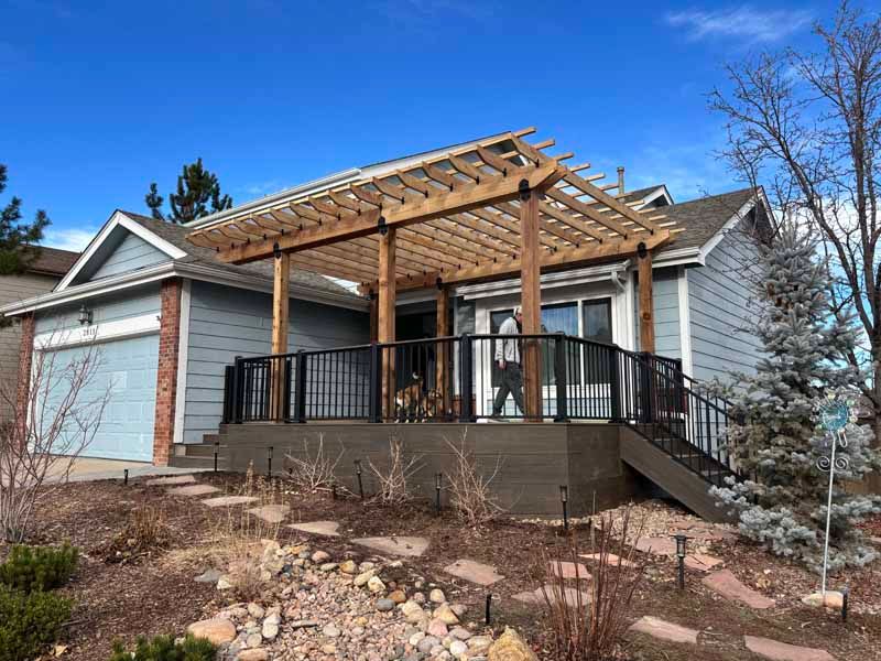 A light blue house features a wooden pergola over a dark-colored deck with black railings and stairs.