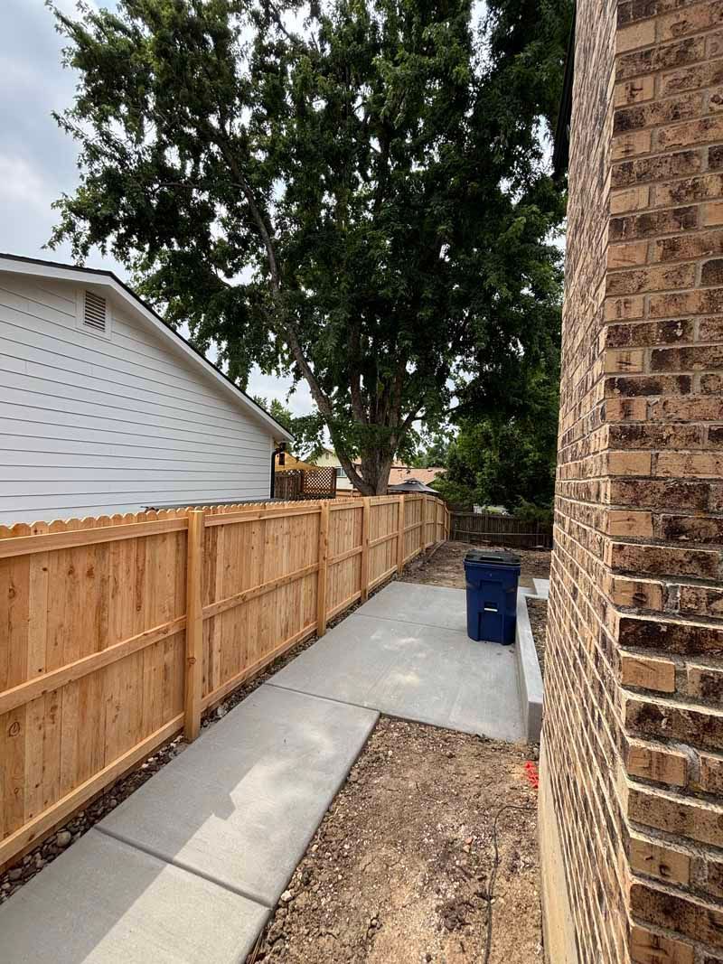 A sidewalk runs between a wooden fence and a brick house, leading to a blue trash bin under a large tree.