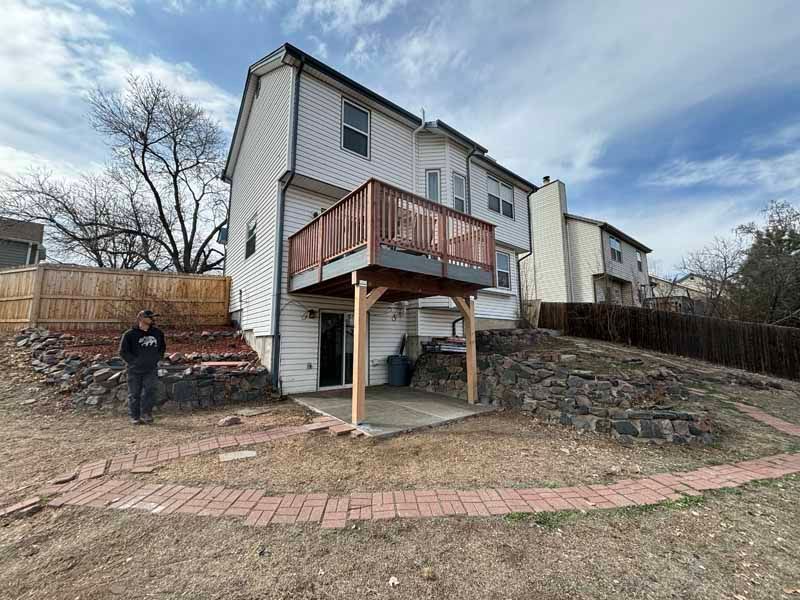 A person stands in a backyard near a light-colored two-story house with a raised wooden deck and a brick pathway.