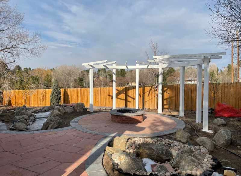A circular brick patio with a fire pit under a white pergola in a backyard with a wooden fence and rocky landscape.