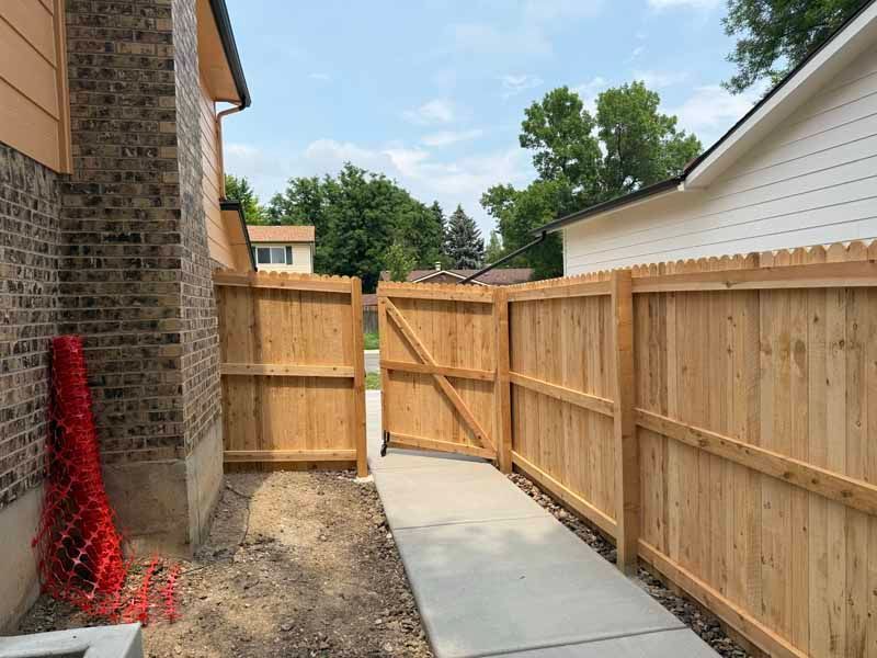 A concrete walkway leading to an open wooden gate between a brick house and a backyard fence.