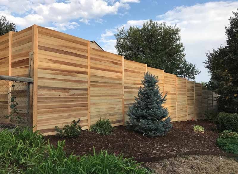 A tall, horizontal-plank cedar fence steps down a gentle slope in a mulched garden with trees under a partly cloudy sky.