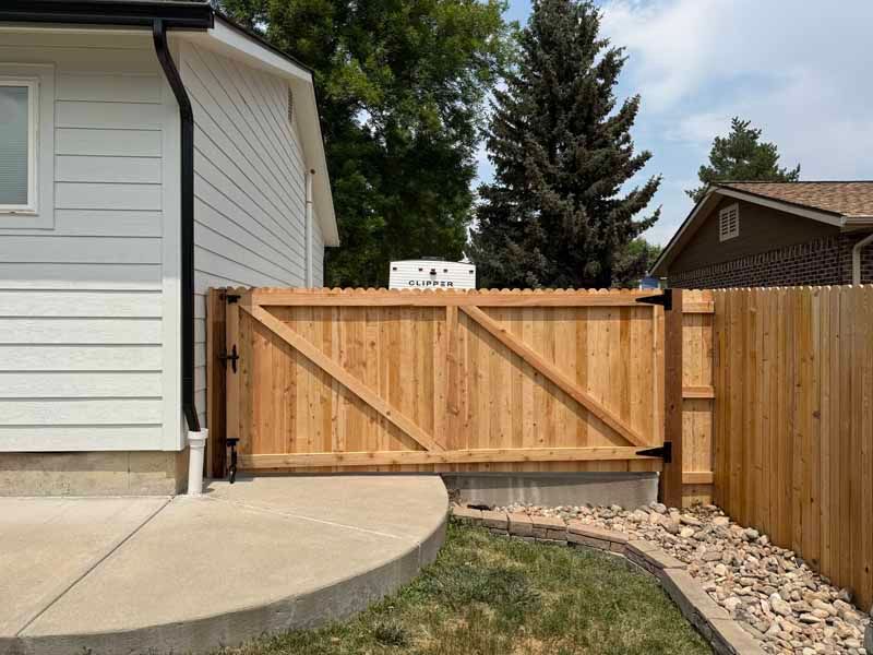 A new wooden gate installed between the white siding of a house and a wooden fence, with a curved concrete patio.