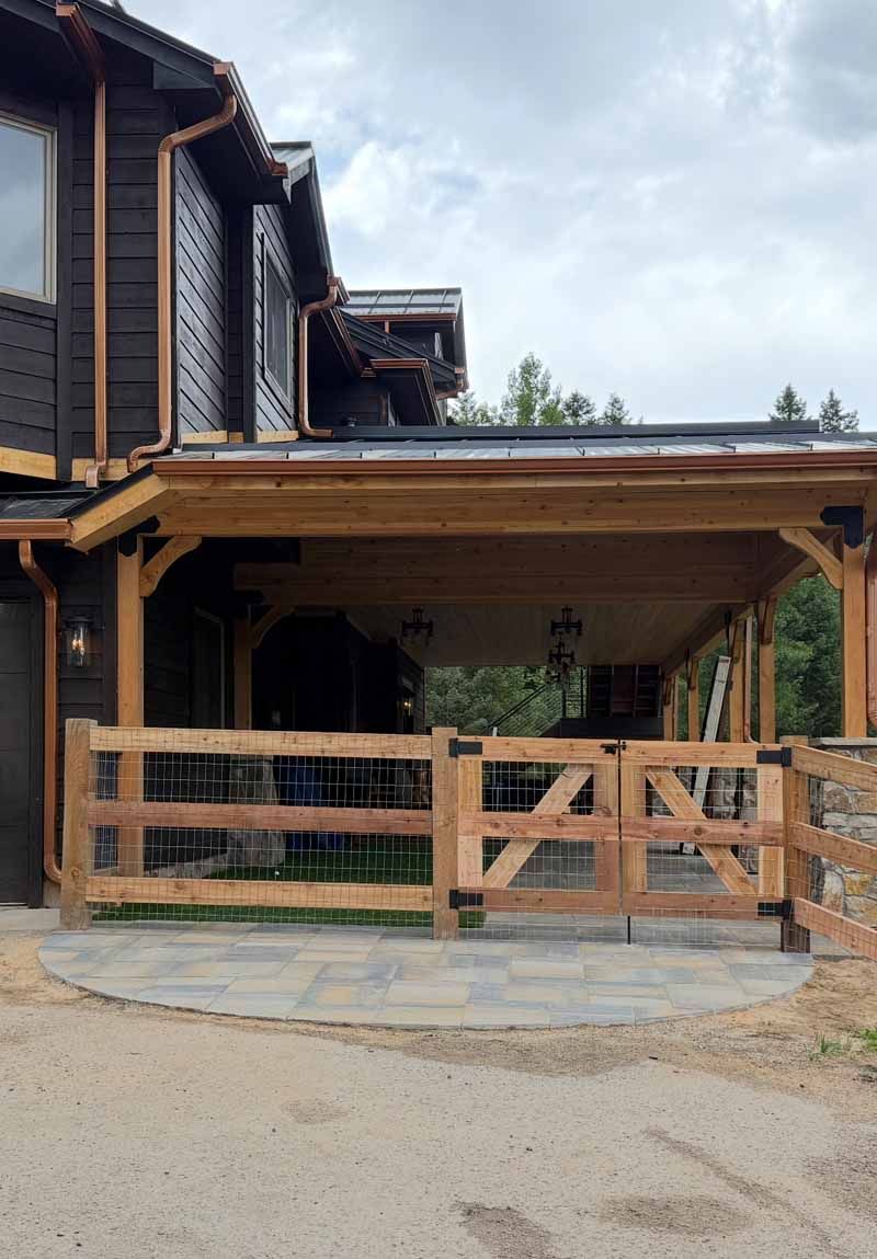 A rustic, dark-wood home with a covered porch and stone patio, enclosed by a wooden fence with a matching gate.