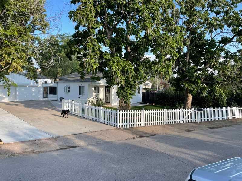 A white, single-story house with a white picket fence, a driveway, and a dog standing on the concrete.