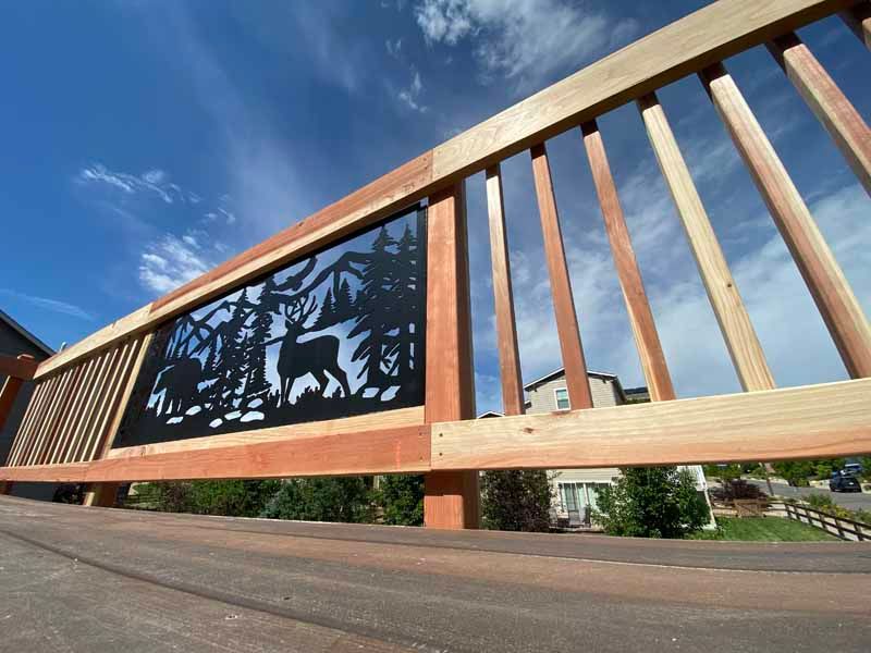 A wooden deck railing features a decorative black metal panel with a cut-out silhouette of a deer in a forest setting.