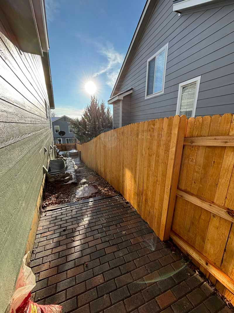 A side yard with a brick pathway running between a house wall and a new wooden privacy fence under a bright sun.