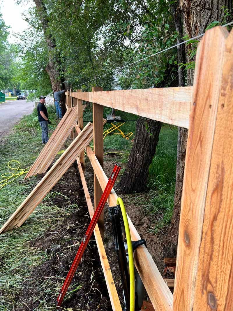 Two workers constructing a wooden fence along a tree-lined street near a road.