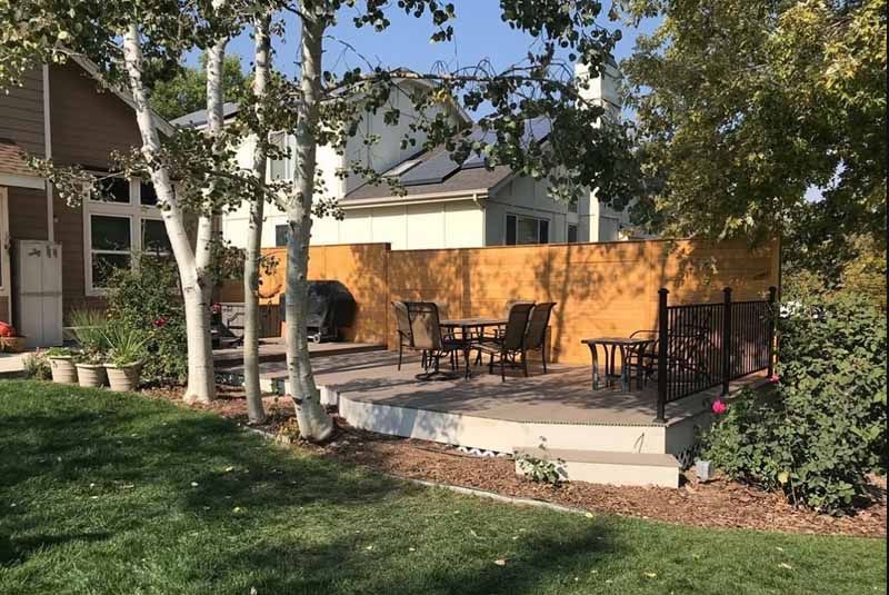 A wooden patio with outdoor furniture, chairs, and a grill sits in a sunny backyard next to birch trees and a house.