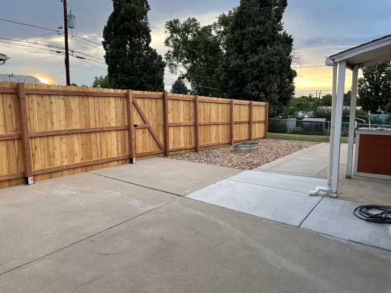 A wooden privacy fence borders a concrete patio and a gravel area under a sunset sky.