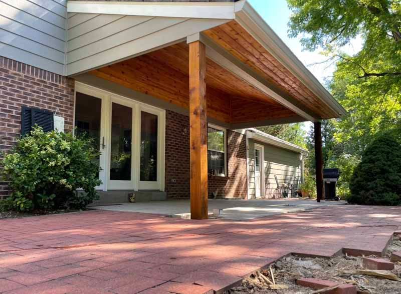 A red brick patio leads to a house with a wooden covered porch featuring a natural wood support post.