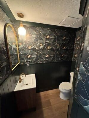 A small bathroom featuring dark floral wallpaper, black paneled lower walls, a wooden vanity, and a toilet.