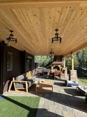 A spacious outdoor patio with a wooden ceiling, stone floor, seating area, and dining space overlooking a wooded yard.
