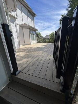 A wooden deck with black railings leads to a house with white siding under a sunny blue sky.