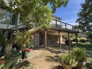 A wooden deck with black railings extends from a stone house into a sunny backyard with trees and potted plants.