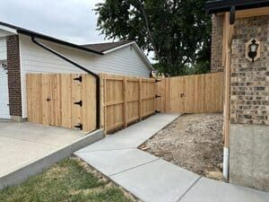 A wooden privacy fence connects a garage to a brick house, bordering a concrete walkway leading to the backyard.