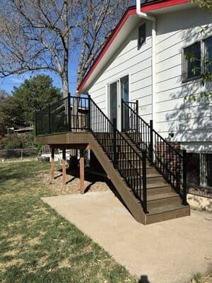A wooden deck with black railings and stairs leading down to a concrete patio outside a white house.