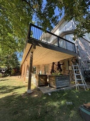Workers build a wooden deck and patio structure attached to the rear of a multi-story house in a backyard setting.