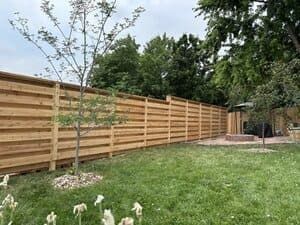 A long, horizontal wooden slat fence spans a backyard with a green lawn, two small trees, and a fire pit area.