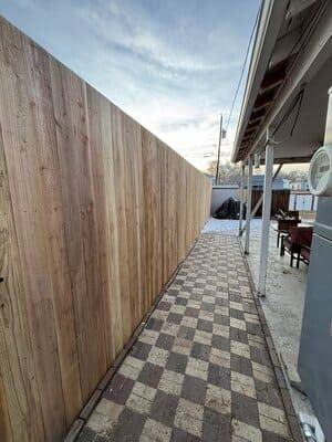 A wooden privacy fence borders a pathway paved with a checkered pattern of light and dark stone tiles.