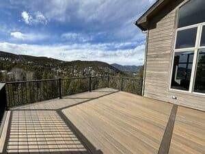 A spacious wooden deck with a black metal railing overlooking a mountain forest landscape under a cloudy blue sky.