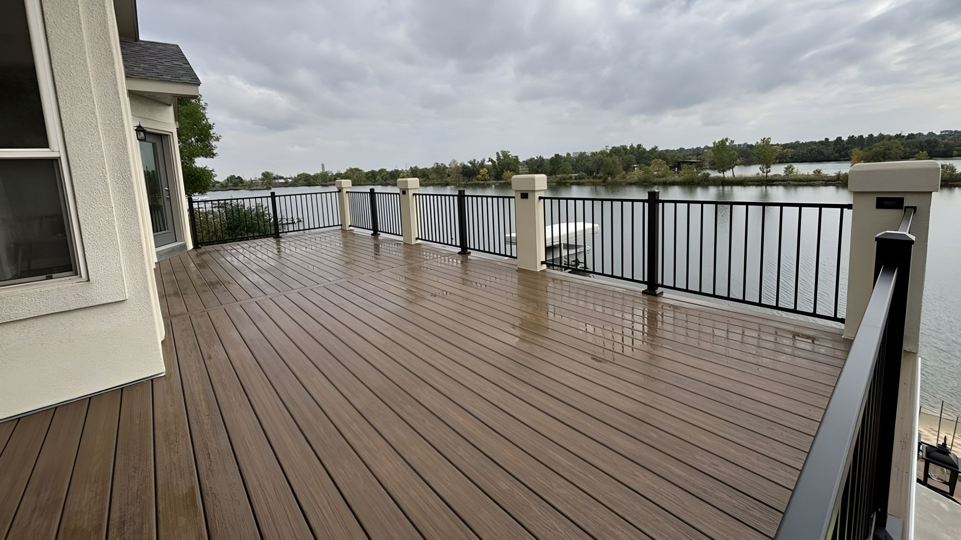 A spacious brown composite deck with black metal railings overlooking a lake under a cloudy sky.
