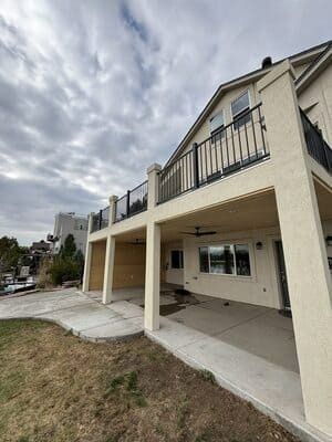 A two-story beige house with a concrete ground-level patio and an upper-level balcony featuring a black metal railing.