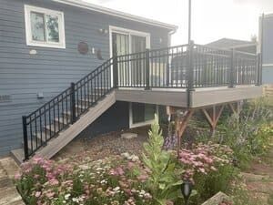 A light blue house with a raised grey deck and black railings, accessed by a set of stairs above a garden bed.