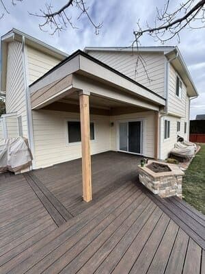 A two-story house with beige siding features a covered deck with dark brown composite planks, a single post, and fire pit.