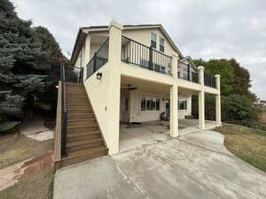 A multi-level home with a tan exterior, wooden deck stairs on the left, a covered patio underneath, and a balcony above.