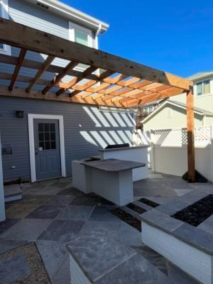 A backyard patio featuring a wooden pergola, outdoor kitchen, stone pavers, and a fire pit under a clear blue sky.