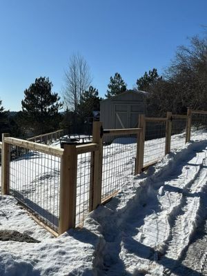 A newly installed wood and wire mesh fence sits on a snowy hillside leading to a storage shed under a clear blue sky.