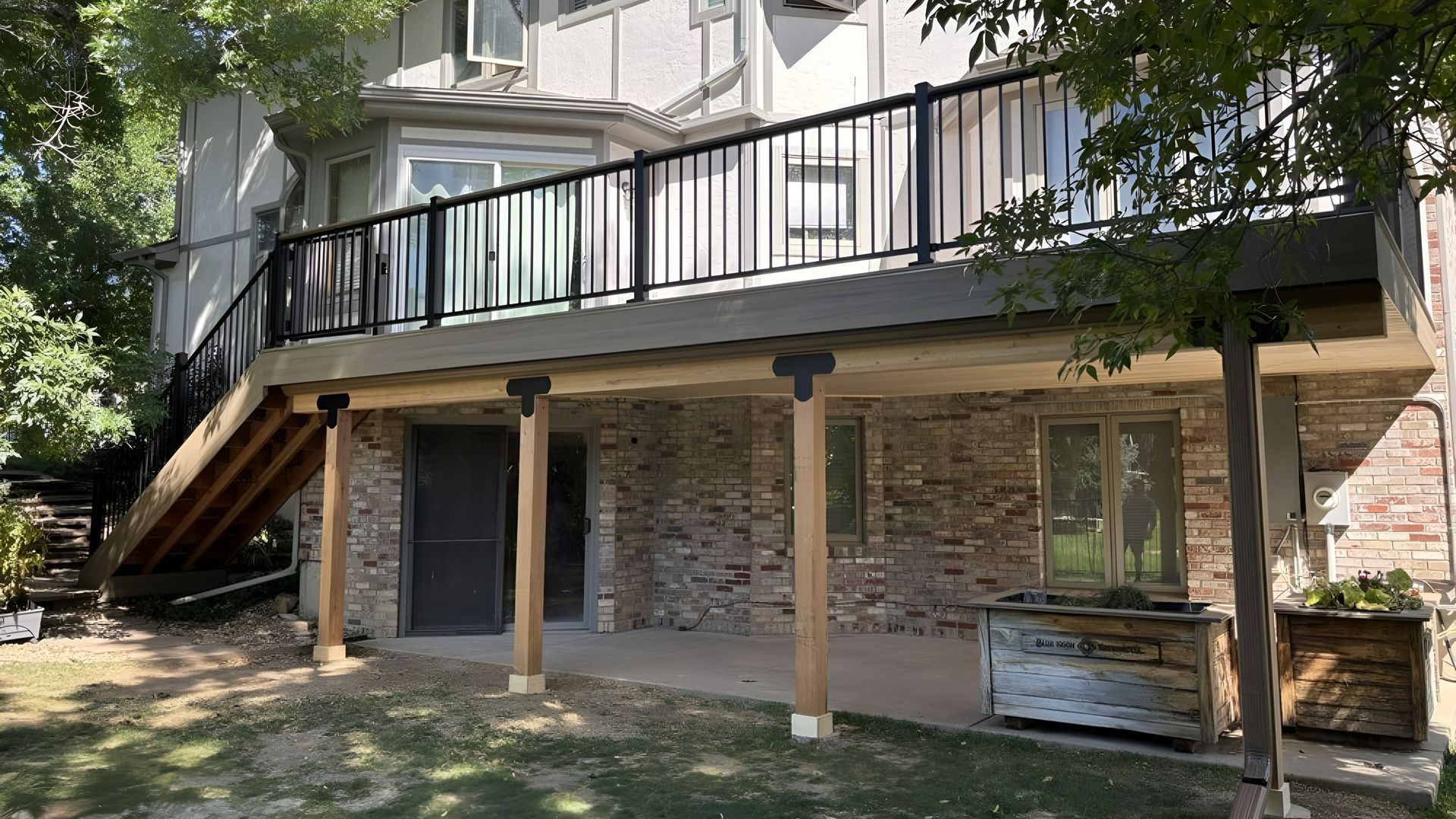 A raised wooden deck with a black railing, featuring stairs leading down to a stone patio area against a house.