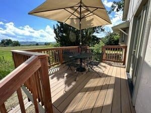 A wooden deck with a table, chairs, and a large umbrella, overlooking a rural landscape with mountains in the distance.